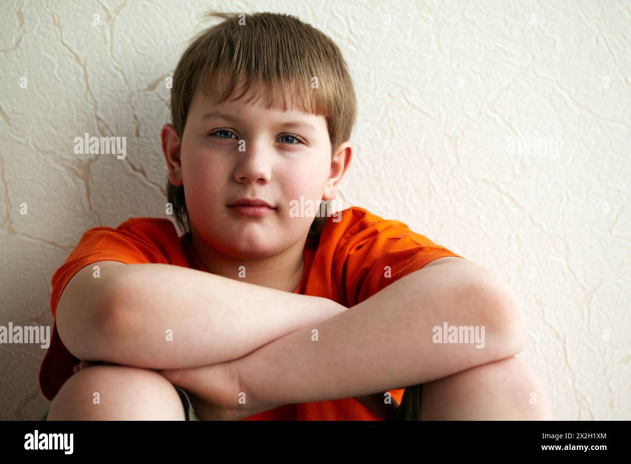 Boy sits with his back to the wall, putting his folded arms on the feet bent in knees Stock Photo