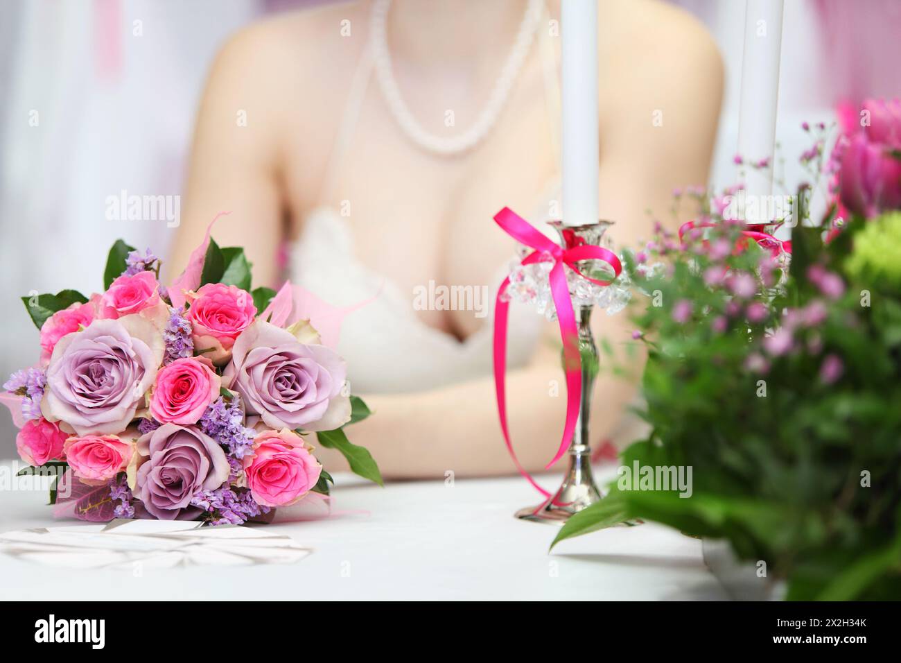 decollete of young bride wearing white dress, which sits at table with candles and holds bouquet of roses; focus on flowers Stock Photo