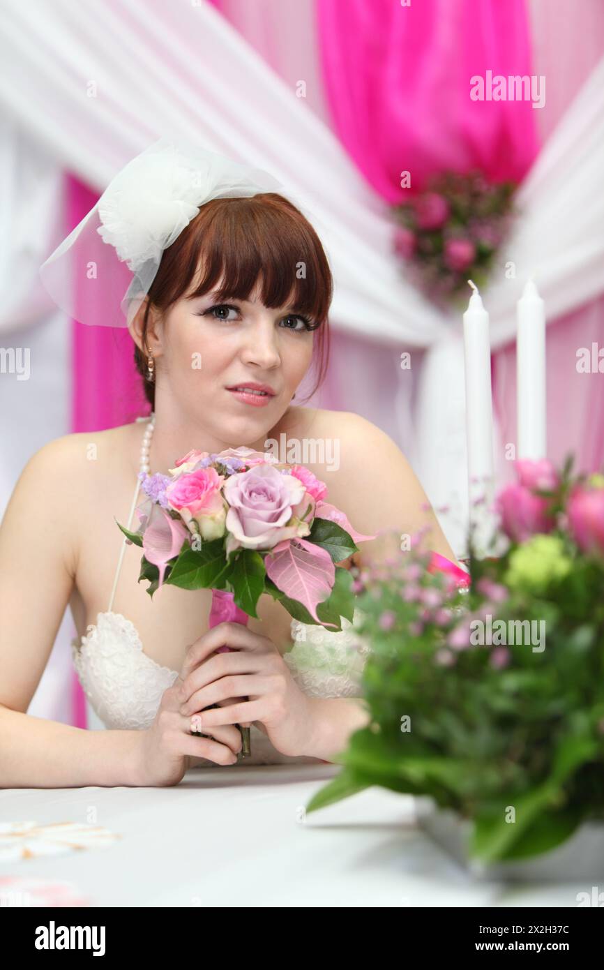 beautiful bride wearing white dress sits at table with candles and looks at camera Stock Photo