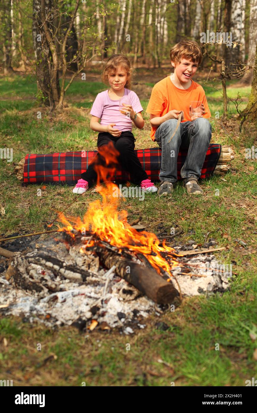 Two children sit near campfire and hold in their hands glasses; focus on boy Stock Photo