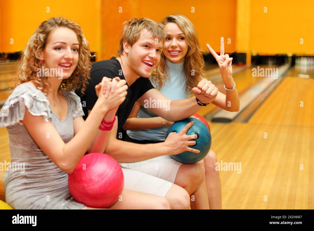 Young man and two happy beautiful girls sit, hold balls and congratulate someone in bowling club; focus on man Stock Photo