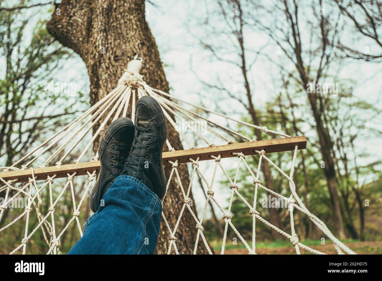 Man laying down and relaxing in back yard hammock attached to a tree, wearing jeans and dirty black sneakers, selective focus Stock Photo
