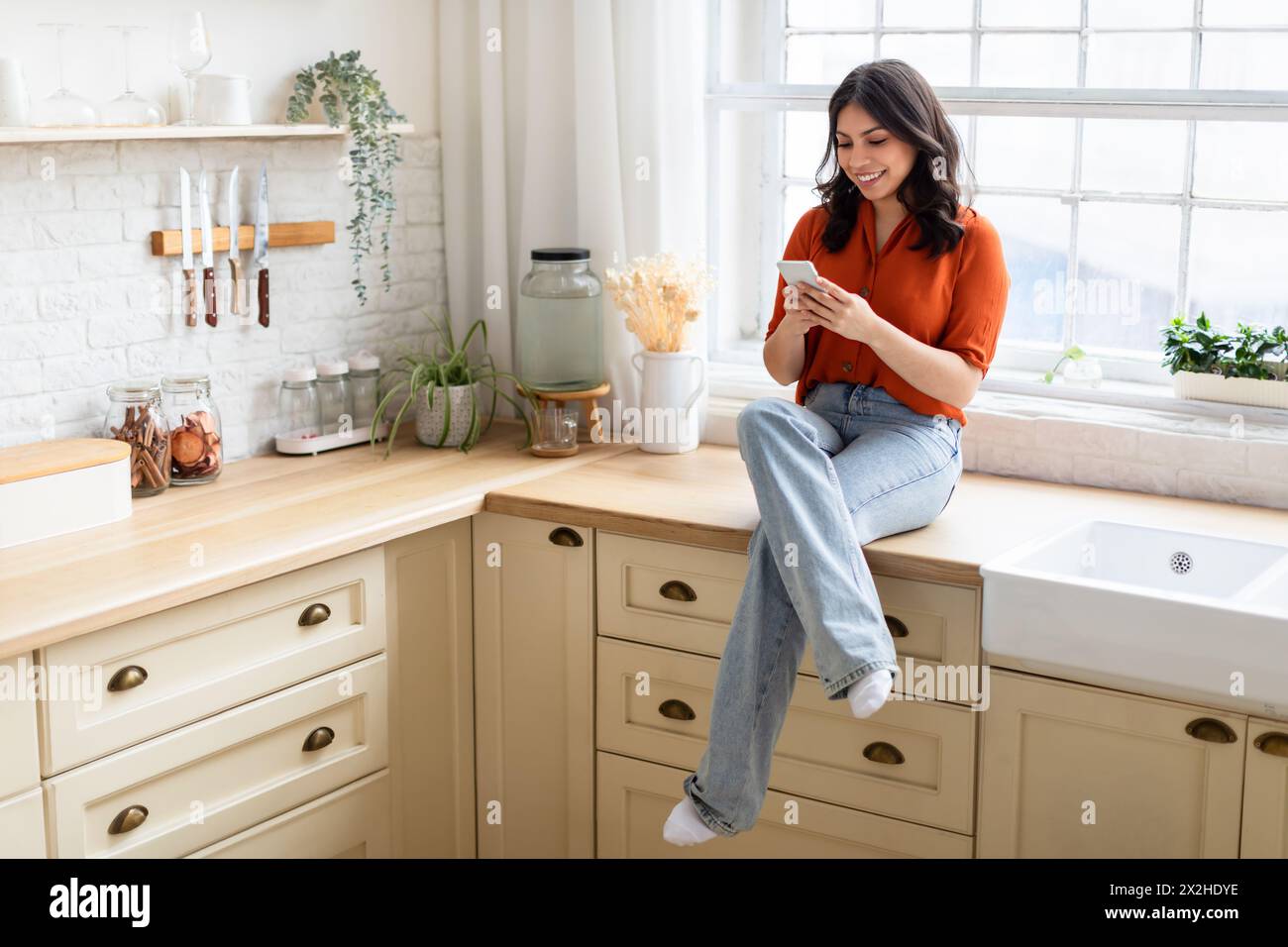 Woman sitting on kitchen counter using smartphone Stock Photo