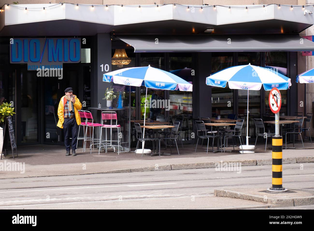 Basel, Switzerland - April 18, 2024: One man standing on the sidewalk in front of restaurant with outdoor seating and umbrellas Stock Photo