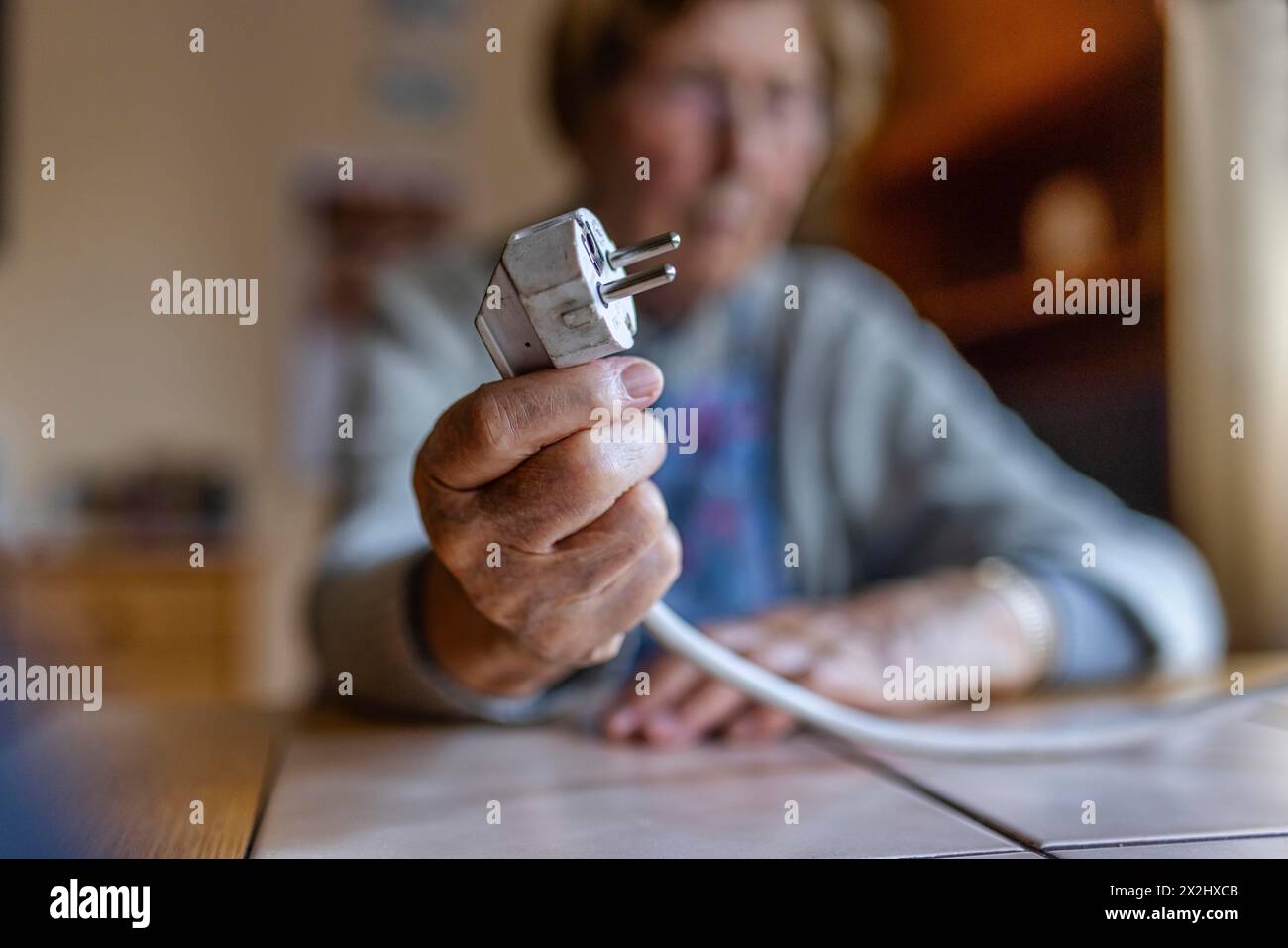 Senior citizen holding a power cable with plug in her hand at home, symbolising energy costs and poverty, Cologne, North Rhine-Westphalia, Germany Stock Photo