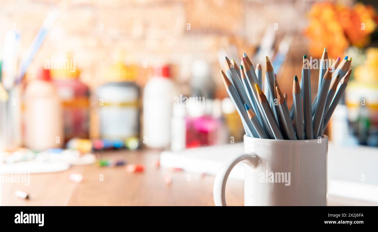 White mug with colored pencils in the foreground on a wooden table with art supplies in the background out of focus Stock Photo