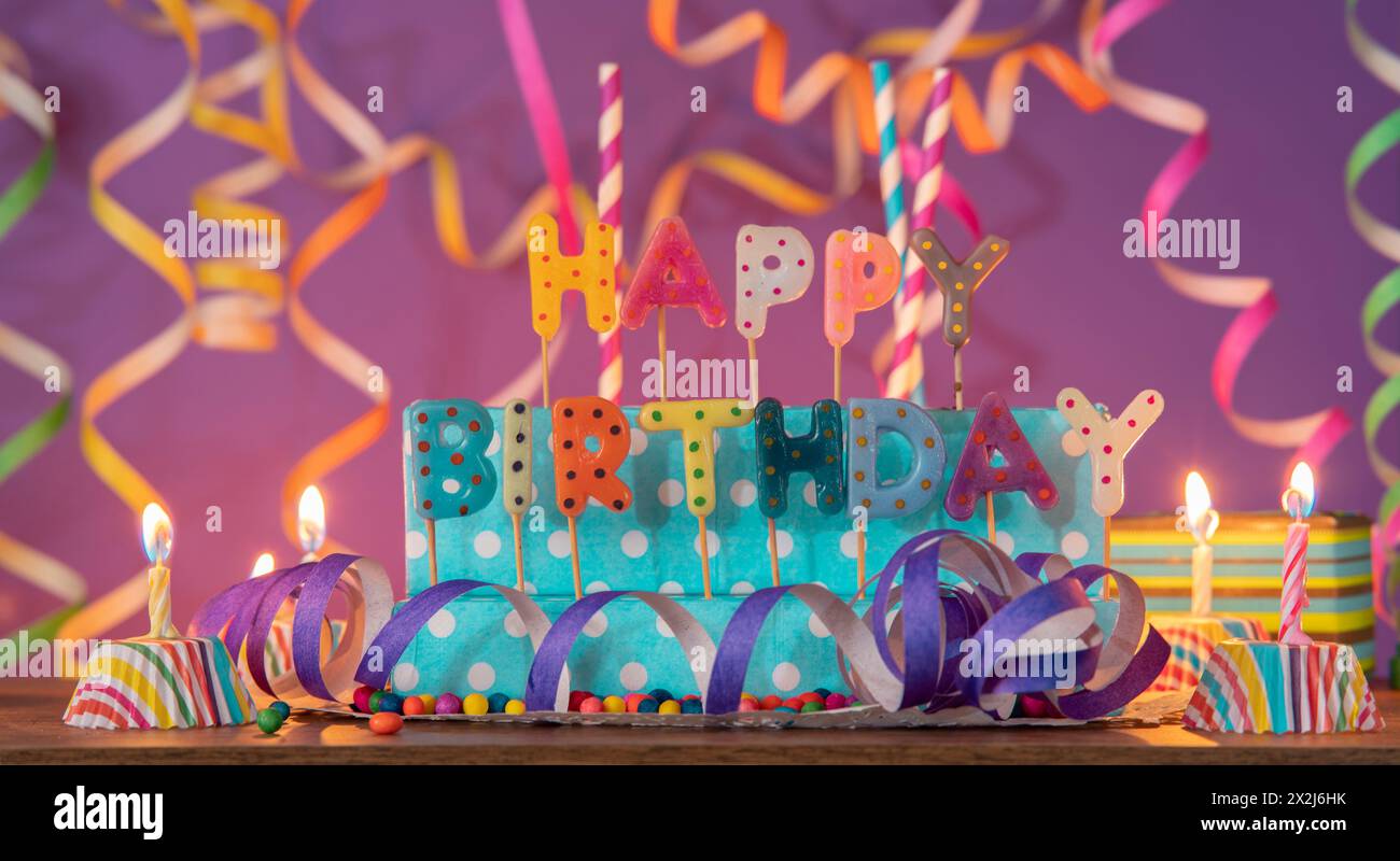 Letters of different colors forming the phrase HAPPY BIRTHDAY, with lit candles against purple out-of-focus background with streamers Stock Photo