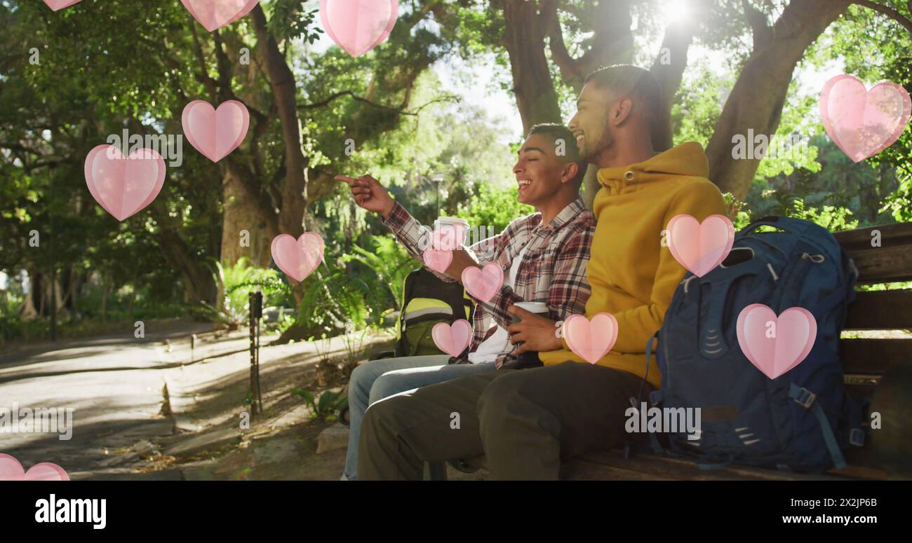 Image of pink hearts floating over happy diverse gay male couple with coffees talking in park Stock Photo