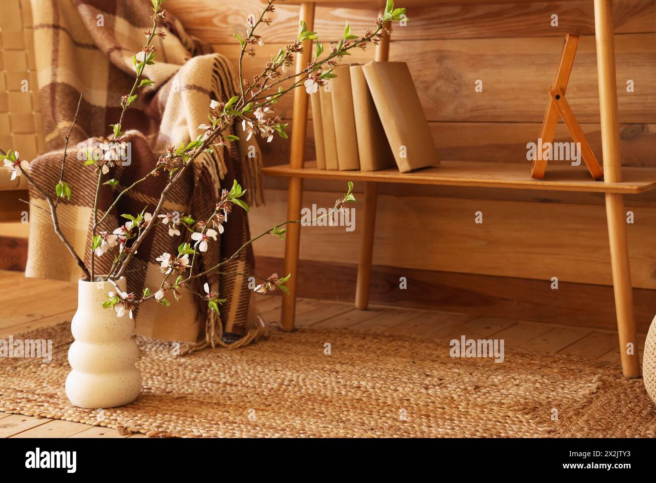 Comfy armchair, shelving unit and vase with blooming branches near wooden wall in living room Stock Photo
