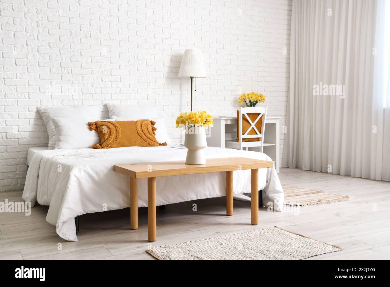 Vase with yellow narcissus flowers on bench near bed in white bedroom Stock Photo