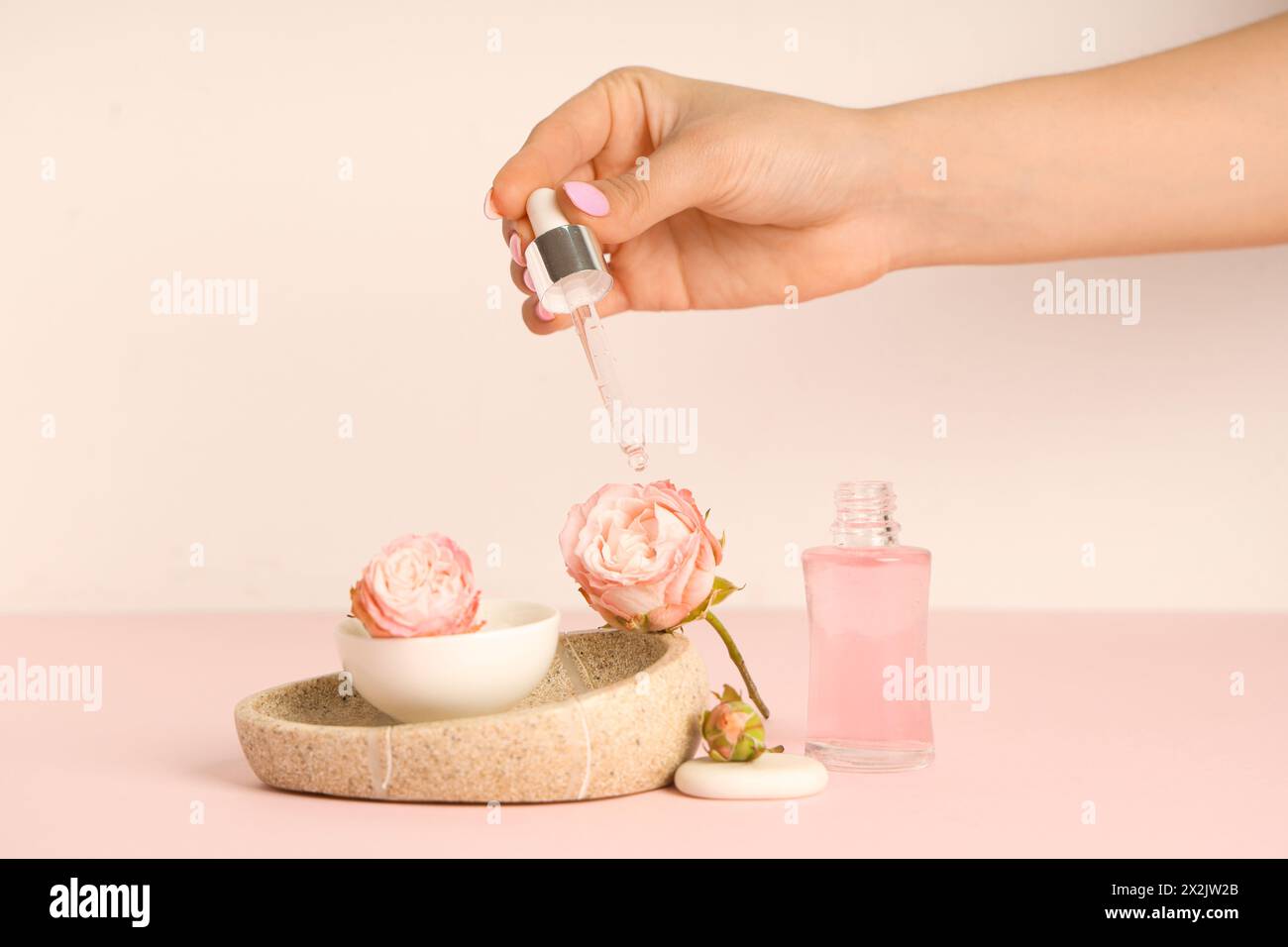 Female hand applying cosmetic oil on roses and stone tray with bowl on pink background Stock Photo