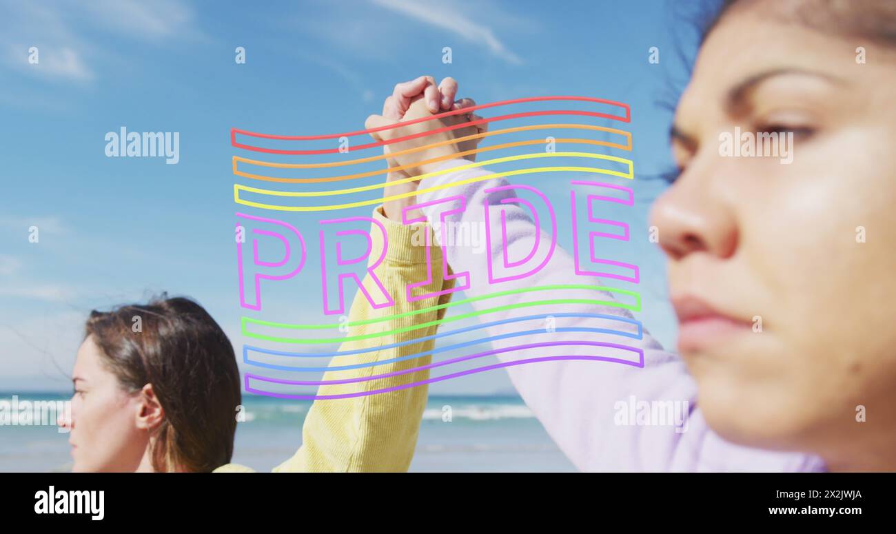Image of pride flag over diverse lesbian couple holding hands by the sea Stock Photo