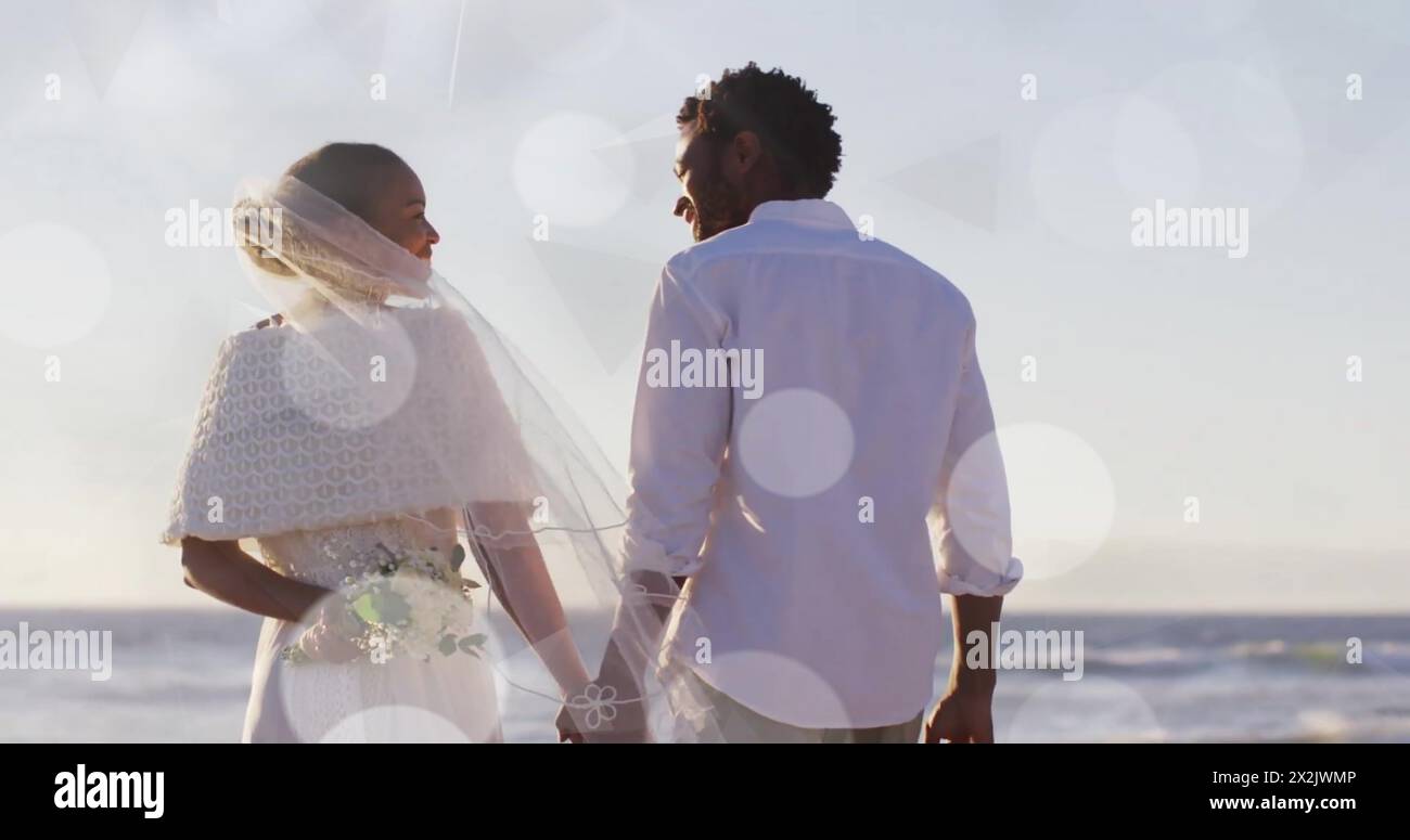 Image of light spots over african american bride and groom holding hands on beach at wedding Stock Photo