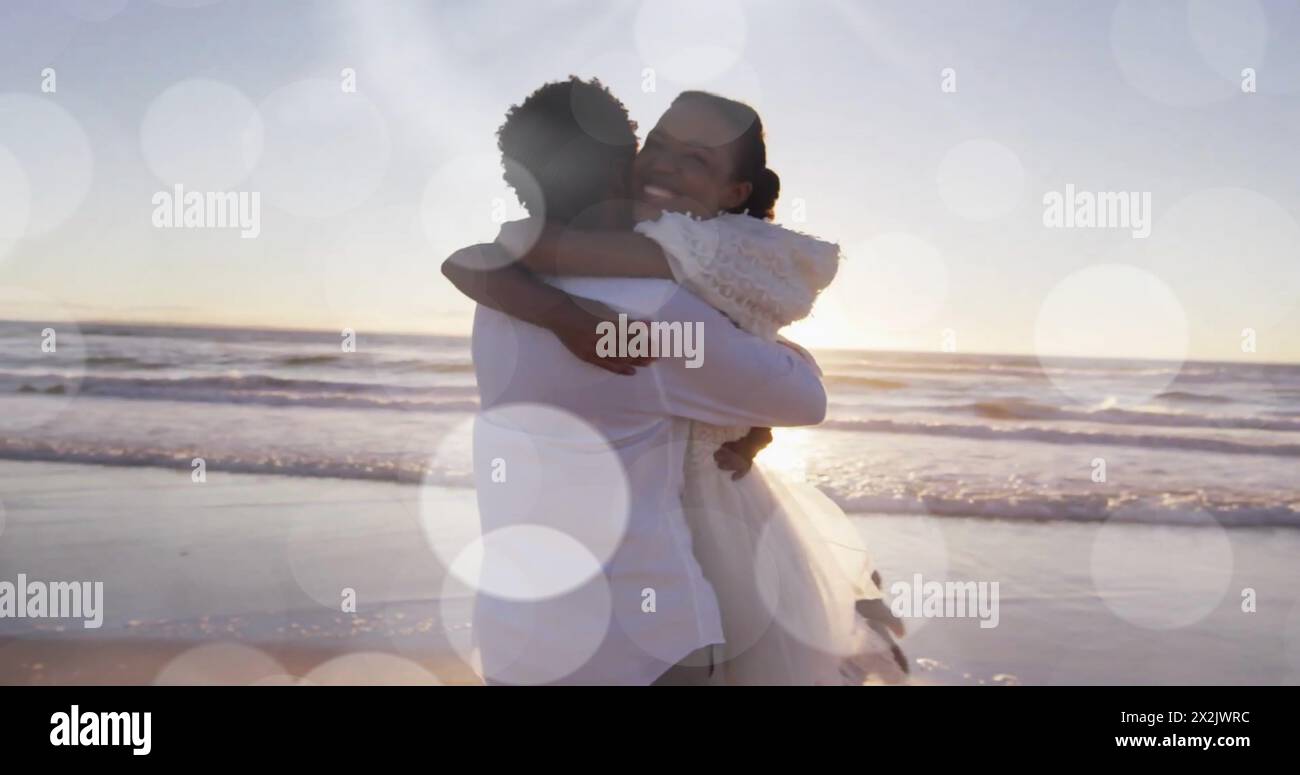Image of light spots over happy african american bride and groom embracing on beach at wedding Stock Photo