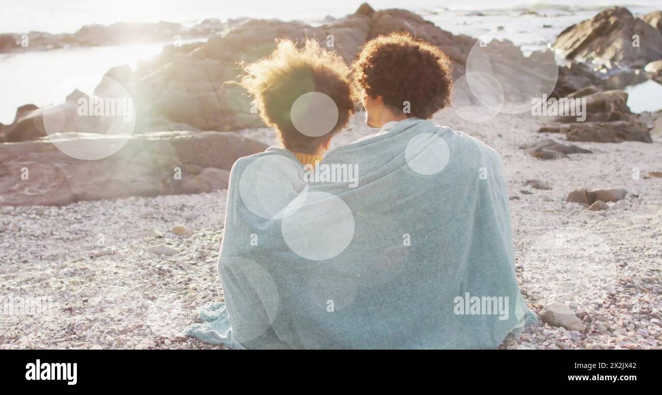 Image of light spots over biracial couple covered in blanket embracing on beach Stock Photo
