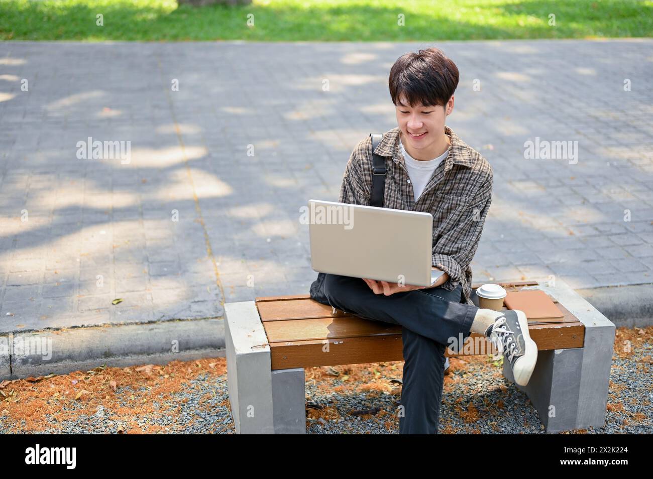 A young Asian male college student using his laptop computer on a bench ...
