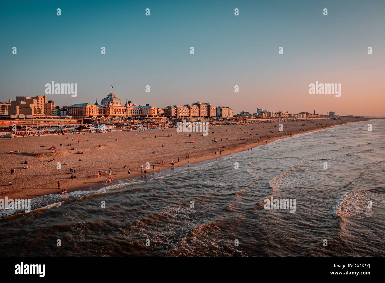 A scenic view of a bustling beach during golden hour, with waves gently lapping the shore and a vibrant city skyline in the background. Stock Photo