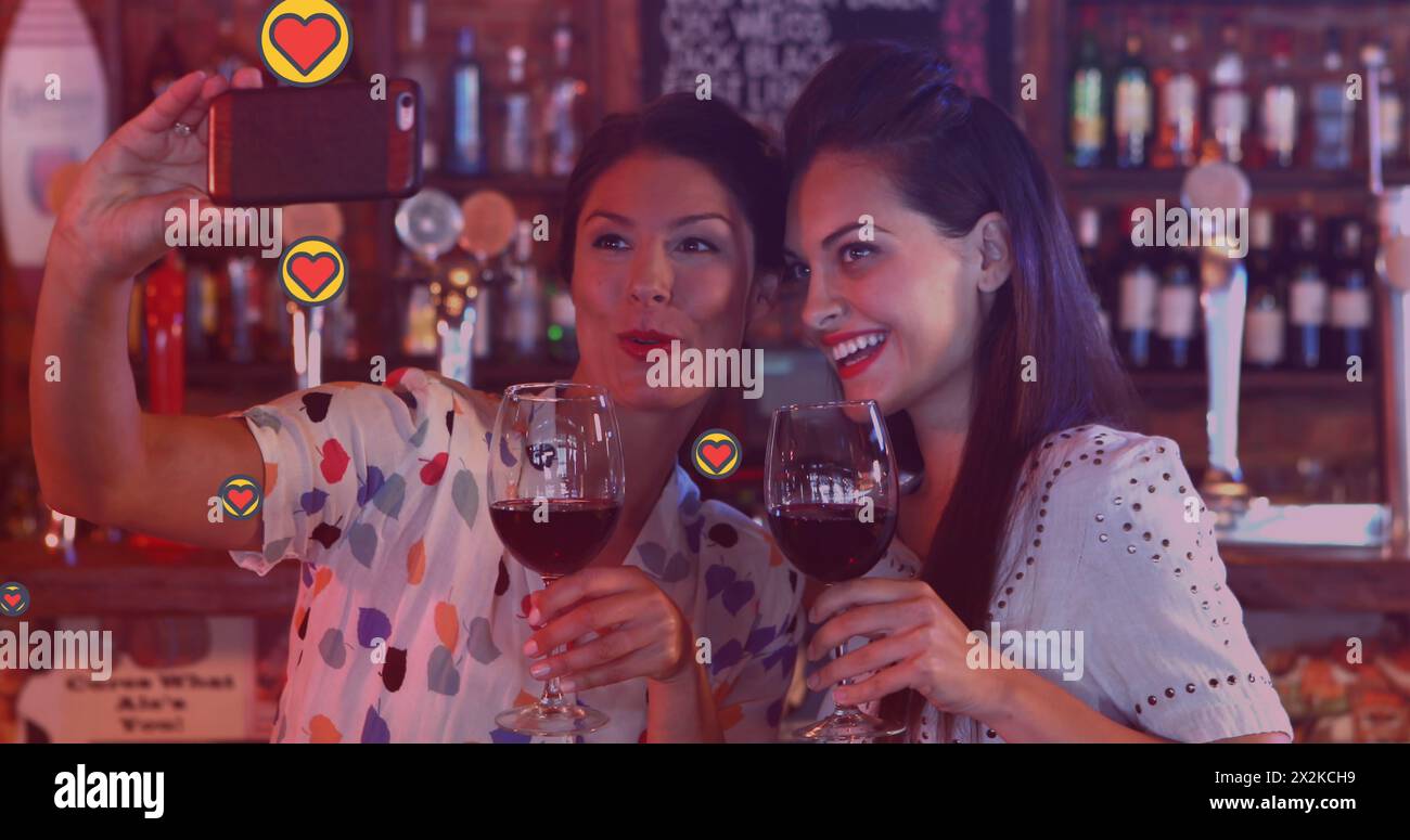 Image of social media reactions over happy caucasian women drinking wine and taking selfie Stock Photo