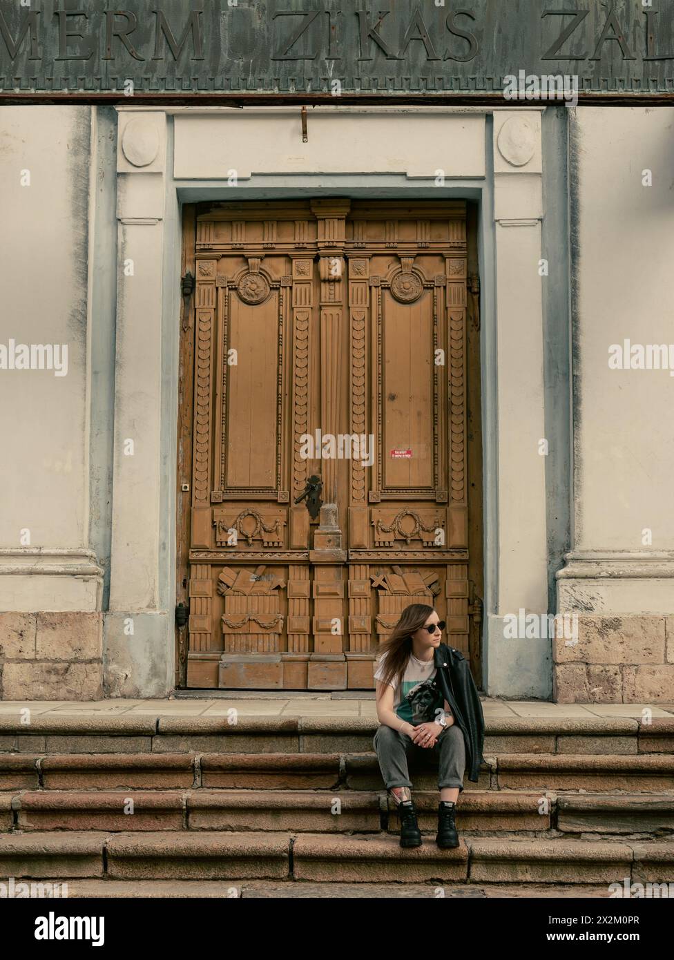 young woman sitting on a steps of “Ave Sol” Concert Hall (previously St. Peter and St. Paul Orthodox Church) with wooden door on a background 28.03.20 Stock Photo