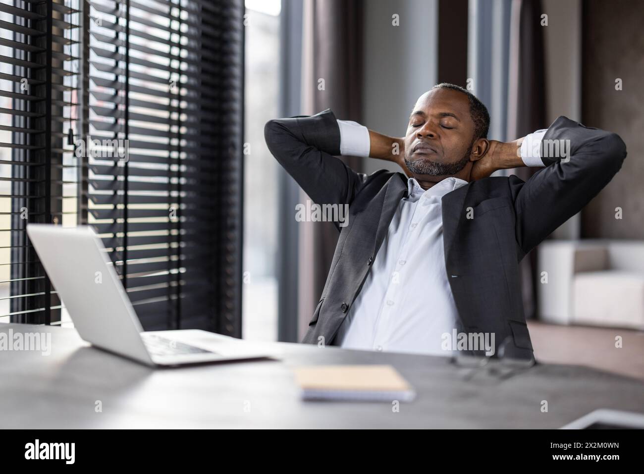 A professional middle-aged black businessman sits back in his office chair with his hands behind his head, eyes closed, embodying relaxation and stress relief amidst a busy work environment. Stock Photo