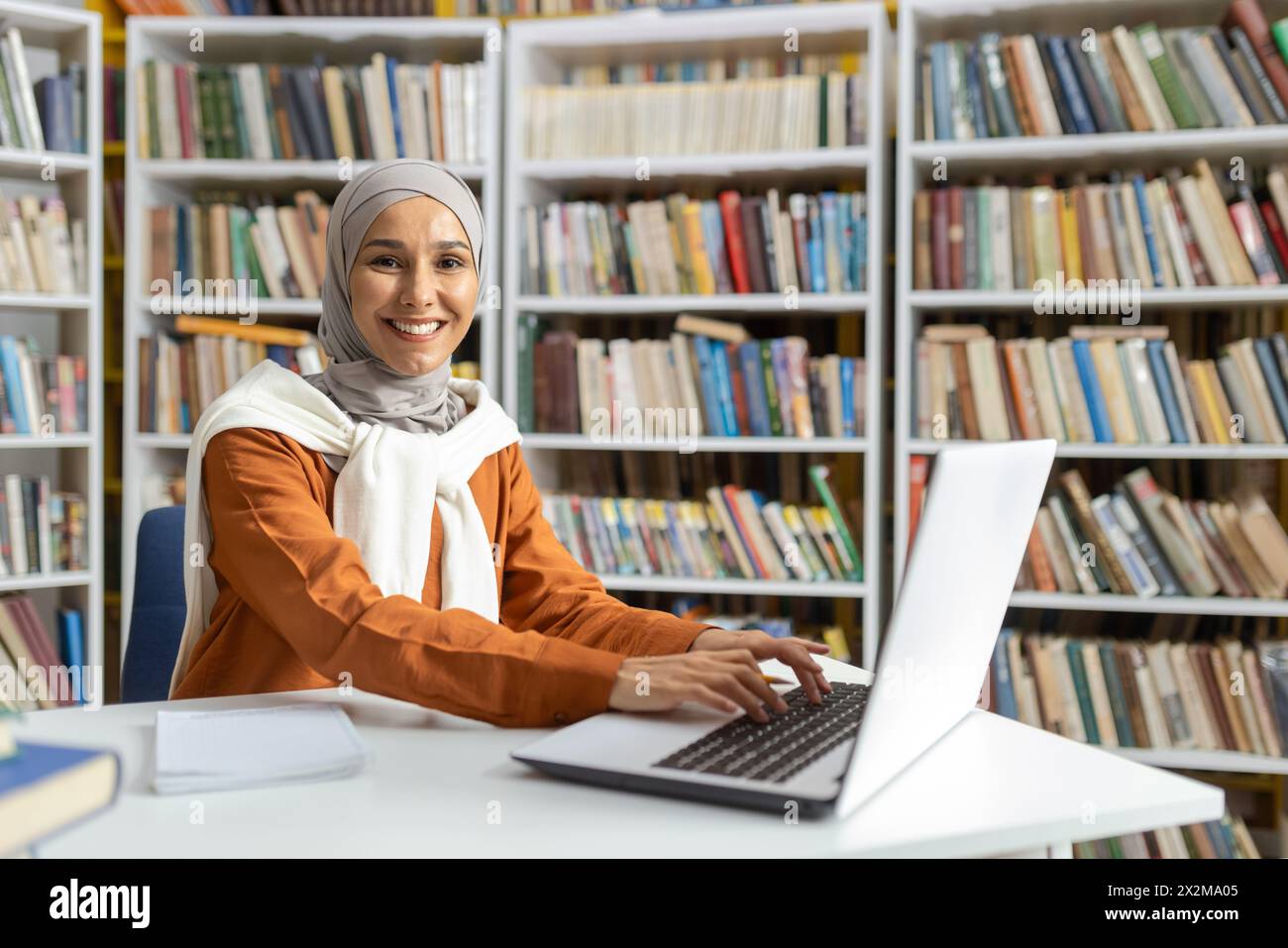 A cheerful Muslim woman in hijab sitting at a desk in a library ...