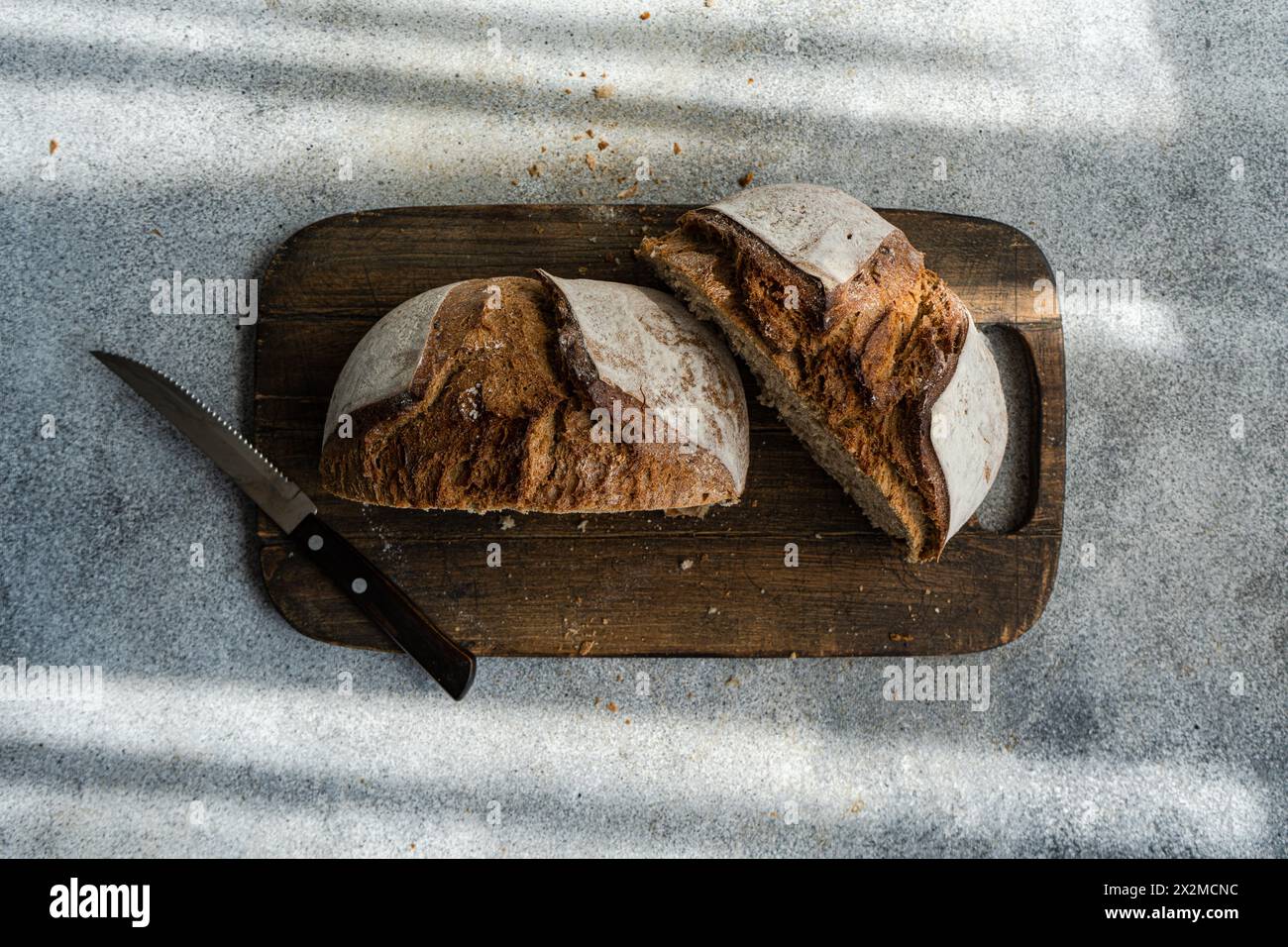 Sliced wholesome rye sourdough bread on a rustic wooden cutting board next to a serrated knife, with natural light casting shadows Stock Photo