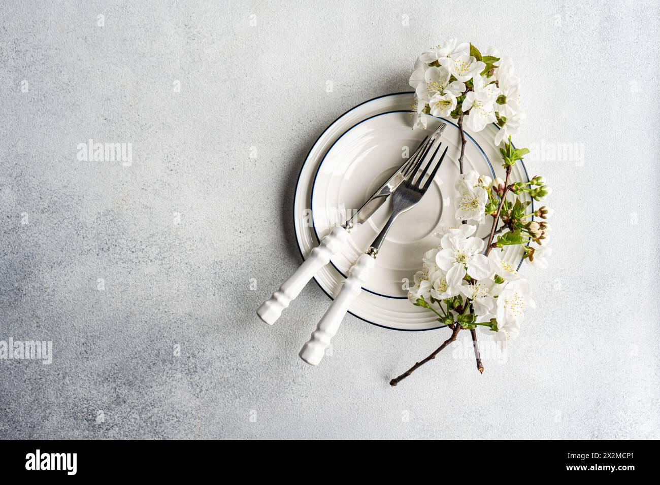 Elegant spring table setting with white blossom branches and vintage-style cutlery on a textured grey background Stock Photo