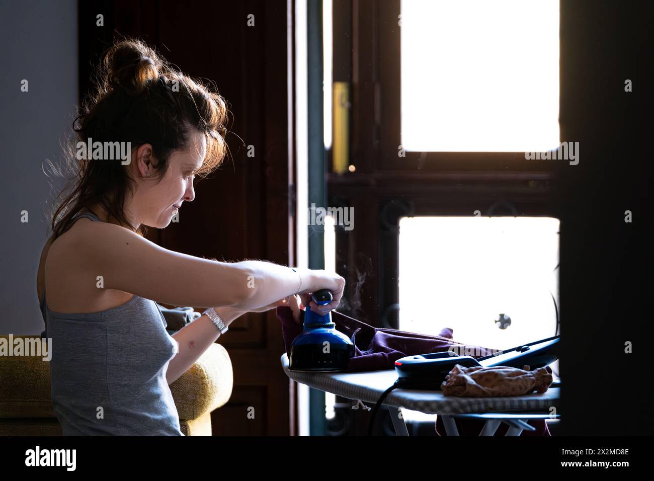 A young woman is focused on ironing clothes on an ironing board in a room with natural light coming from a window Stock Photo