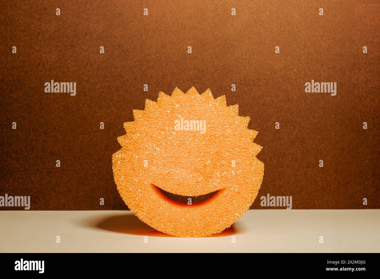 A vibrantly orange sponge with a cut-out smile sits against a brown backdrop, offering a simple yet cheerful visual metaphor for cleanliness and happi Stock Photo