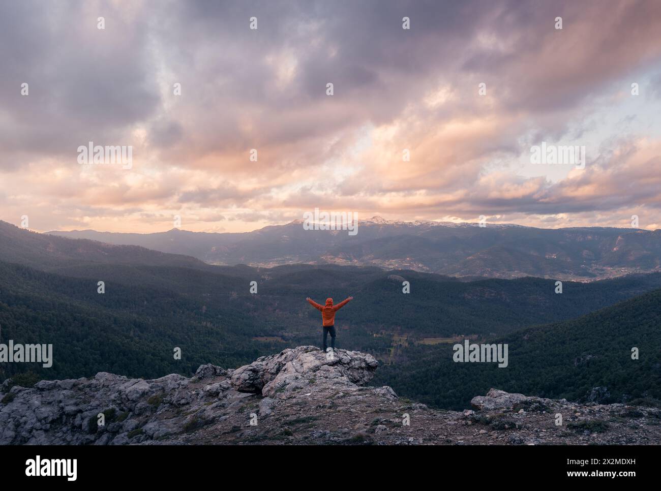 An adventurer stands with arms raised on a rocky peak, celebrating as the sunset paints the skies over the forests and mountains of Rio Mundo in Casti Stock Photo