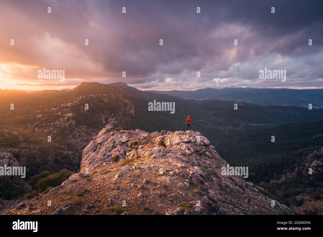 Lone hiker stands atop a mountain at sunset overlooking the vast scenic landscape of Rio Mundo, with dramatic clouds and soft light illuminating the h Stock Photo