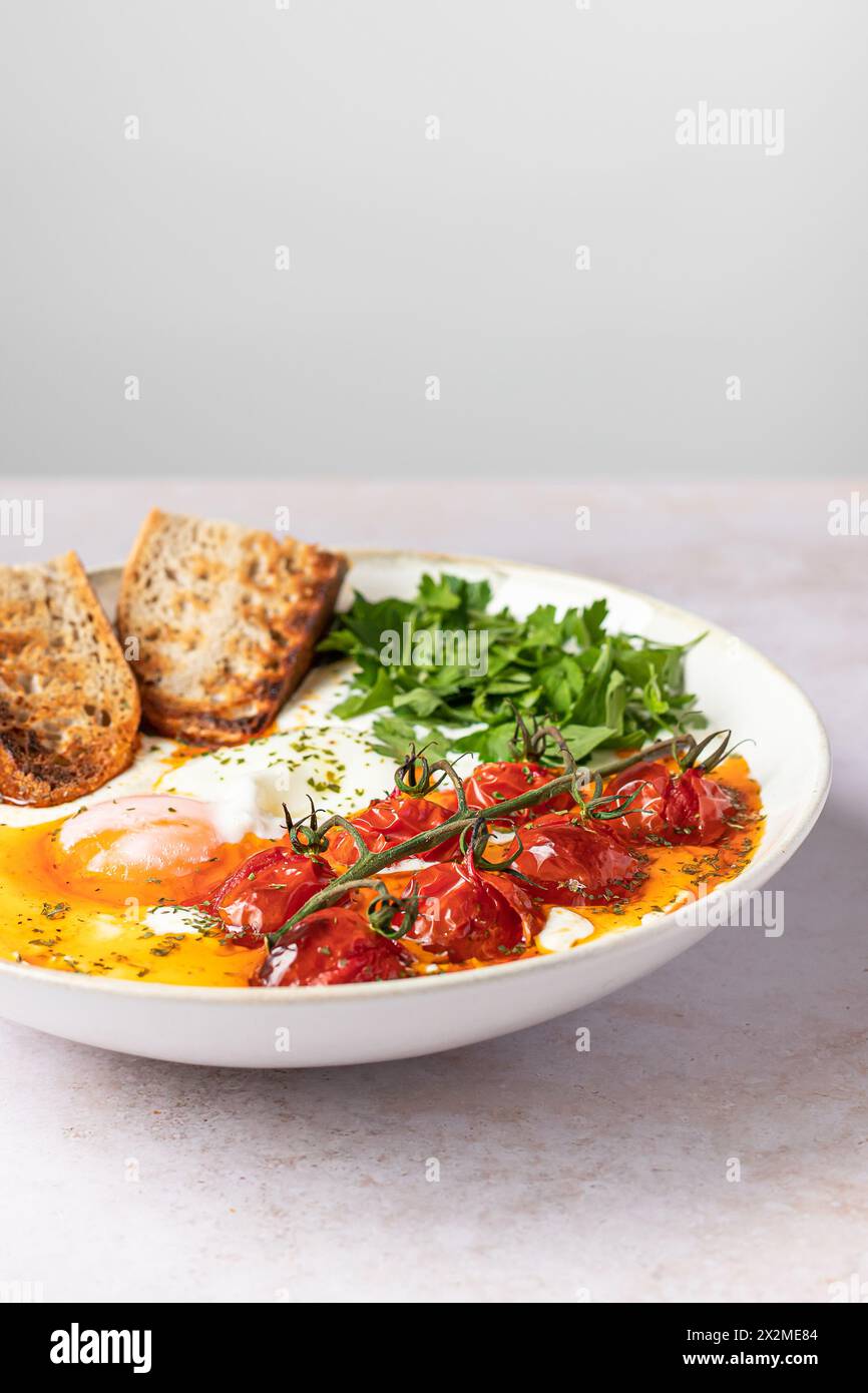 A plate of Turkish Eggs featuring poached eggs on Greek yogurt, topped with spiced butter sauce, cherry tomatoes, and parsley, accompanied by toasted Stock Photo