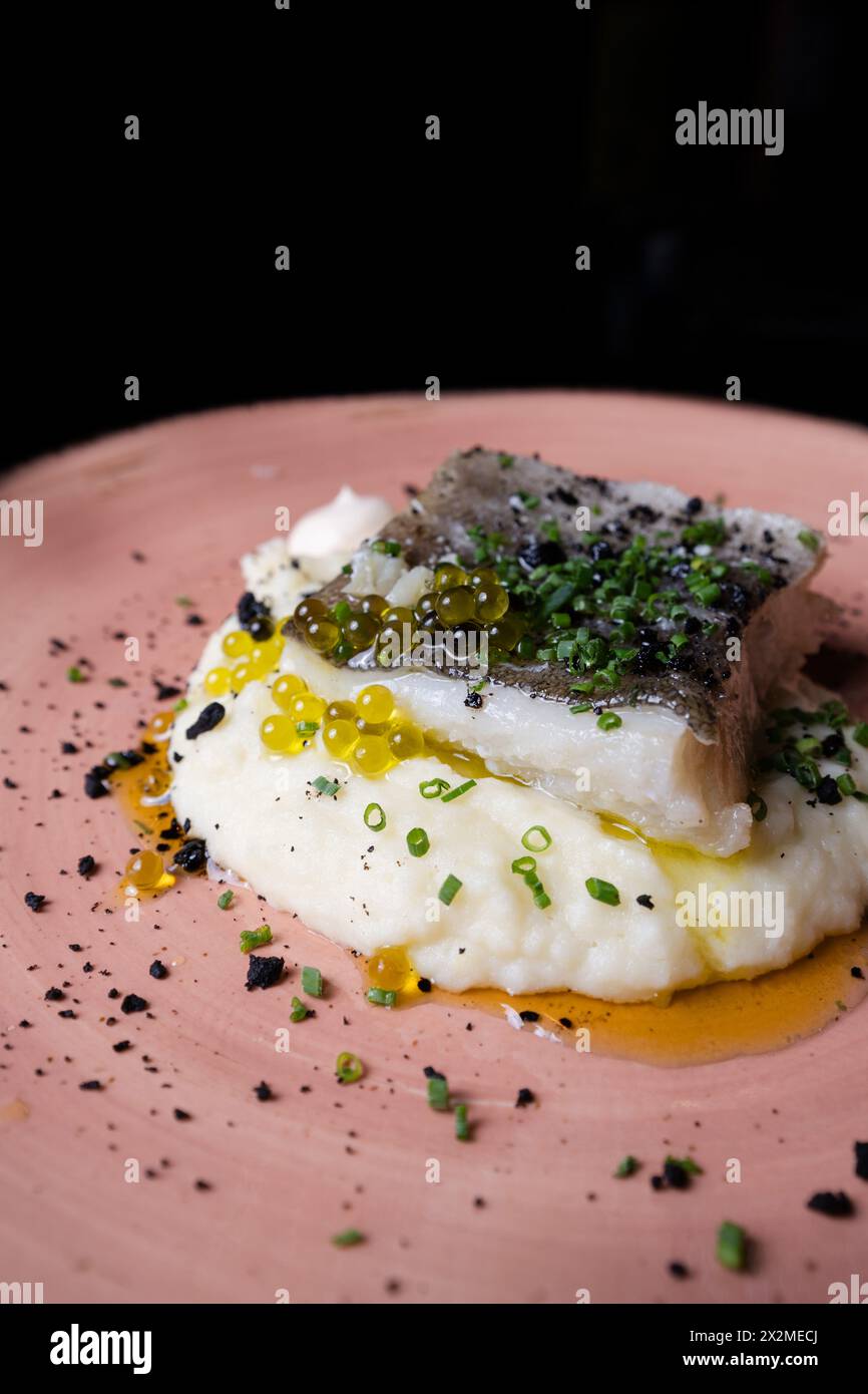 Succulent cod confit laying on a bed of creamy mashed potatoes, garnished with fresh chives, black sesame, and vibrant olive oil pearls, presented on Stock Photo