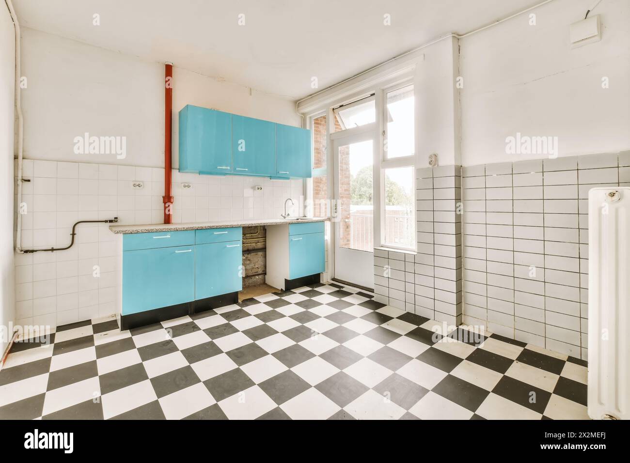 An empty, sunlit kitchen features bright blue cabinetry, white tiles, and a classic black and white checkered floor. Stock Photo
