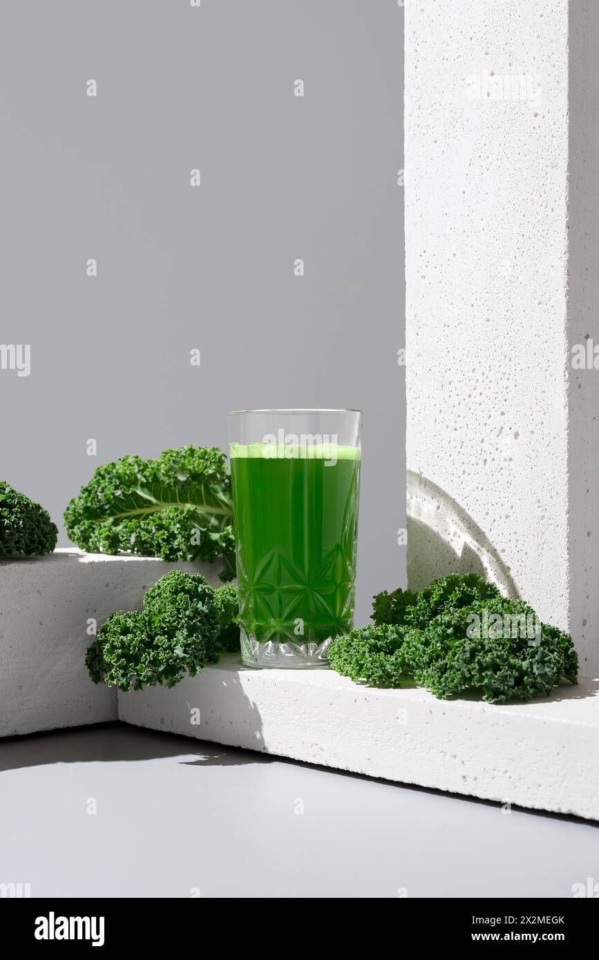 A transparent glass filled with vibrant green kale juice is placed next to fresh, crisp kale leaves on a modern white surface, showcasing a healthful Stock Photo
