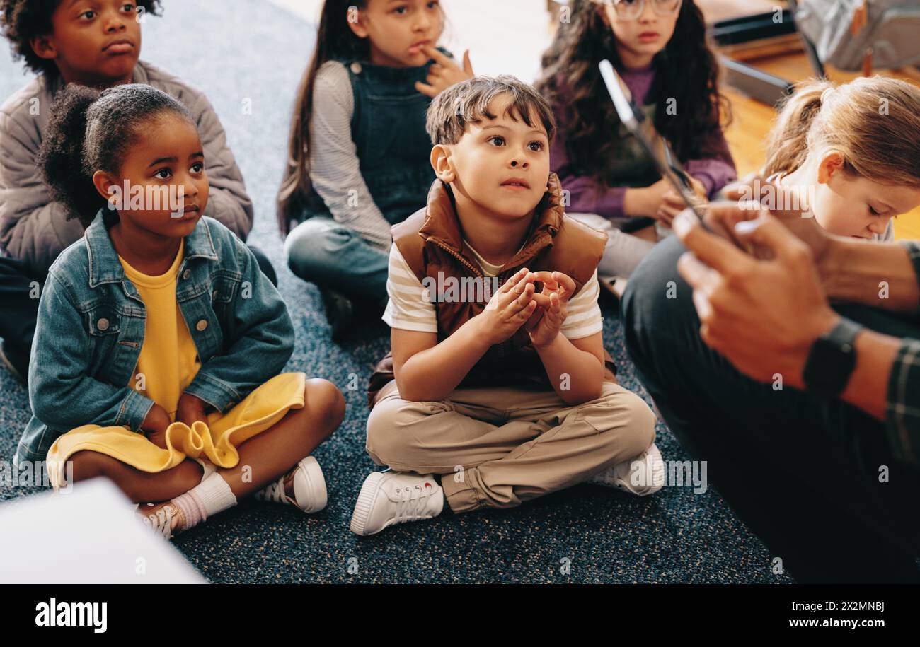 Listening comprehension lesson in an elementary class. Kids pay attention to their teacher reading a story from a tablet. Group of diverse children si Stock Photo