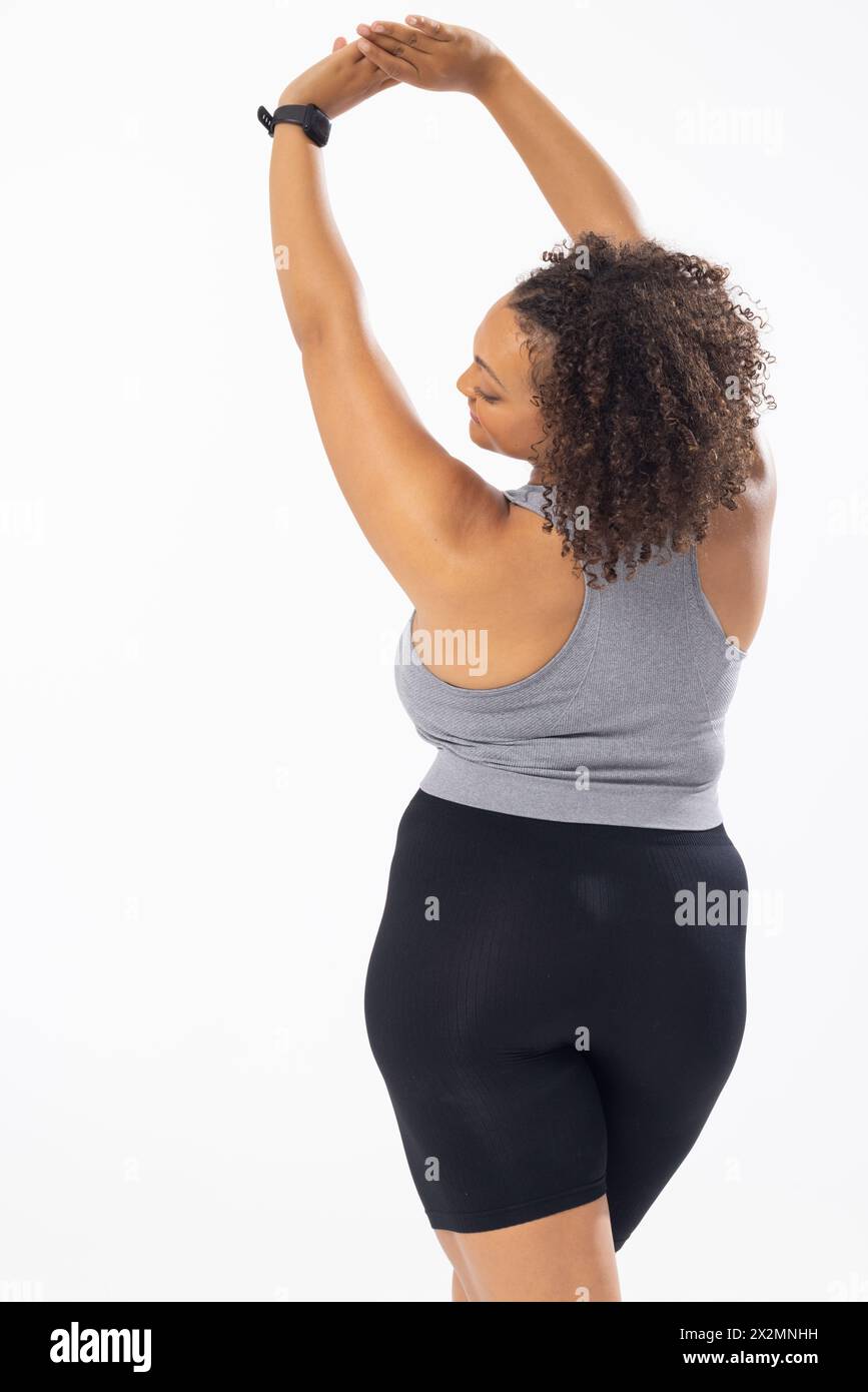 Biracial young female plus size model stretching arms above head on white background, wearing sportswear. She has curly brown hair, light skin, and is Stock Photo