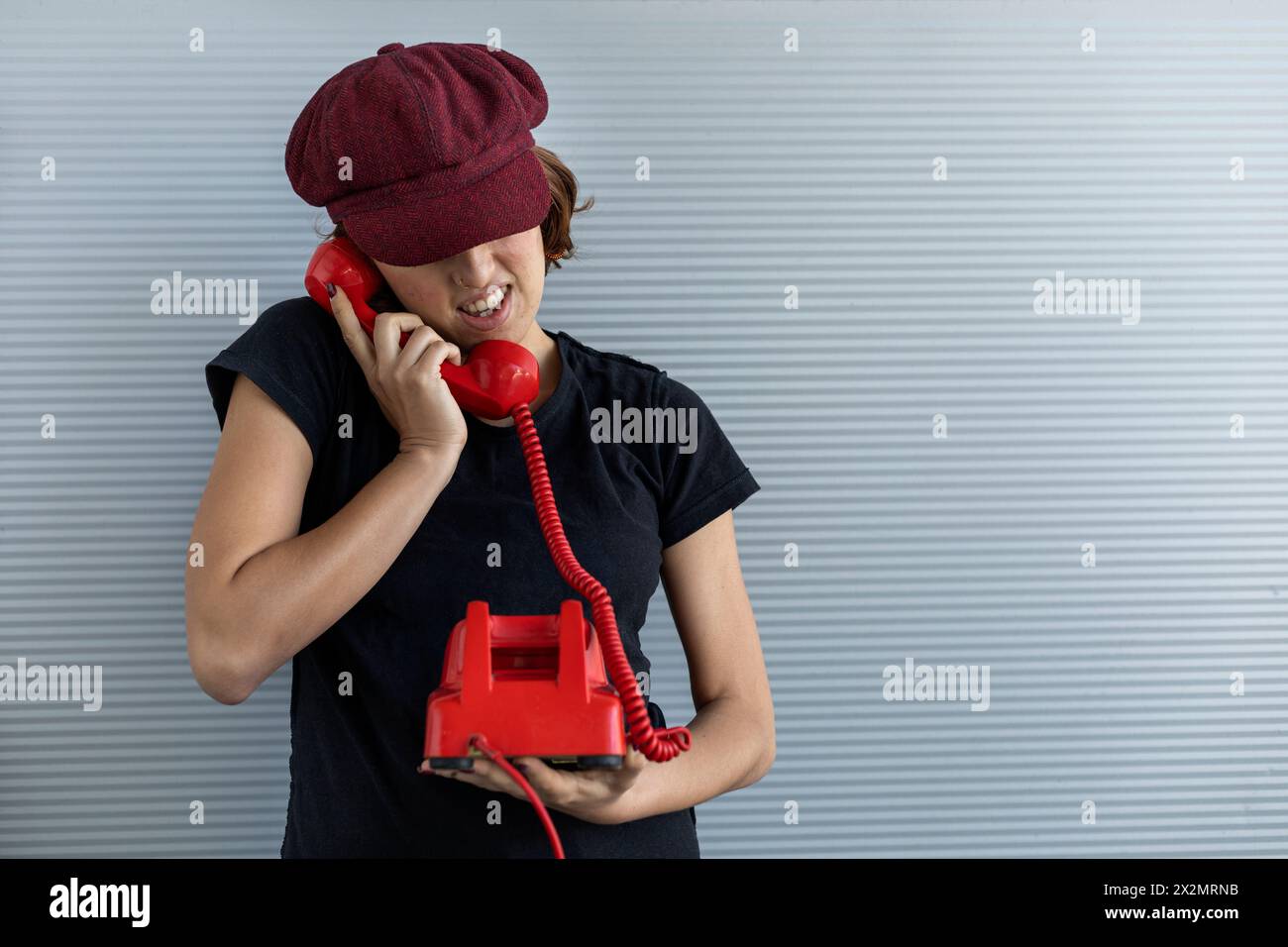 Medium shot of young Latin American girl (22) with cap and red hair talking on retro red phone. Vintage technology concept Stock Photo