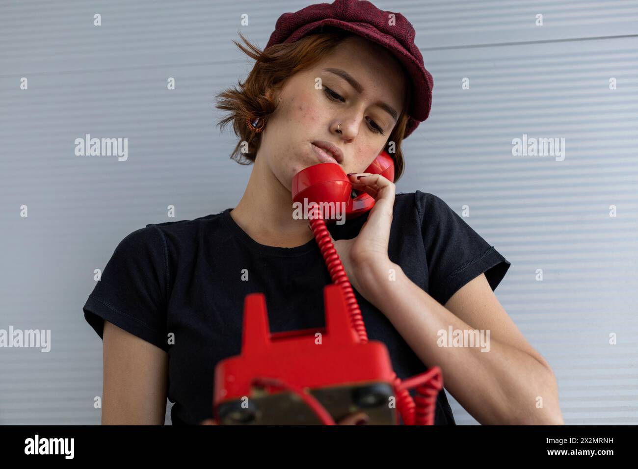 Medium short shot of young Latin American girl (22) with cap and red hair talking on retro red phone. Vintage technology concept. Stock Photo