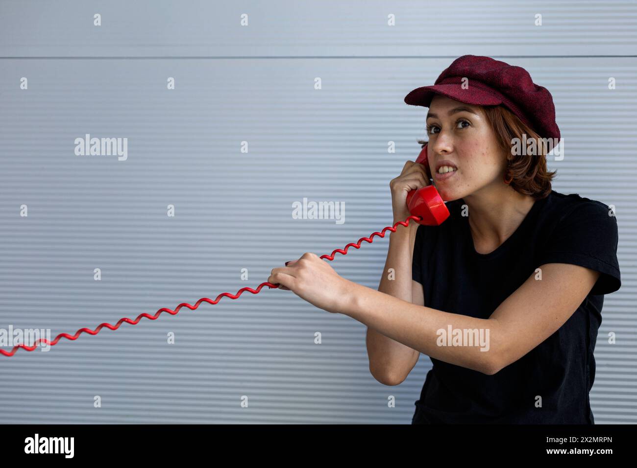 Medium shot of young Latin American girl (22) with cap and red hair talking on retro red phone. Vintage technology concept Stock Photo