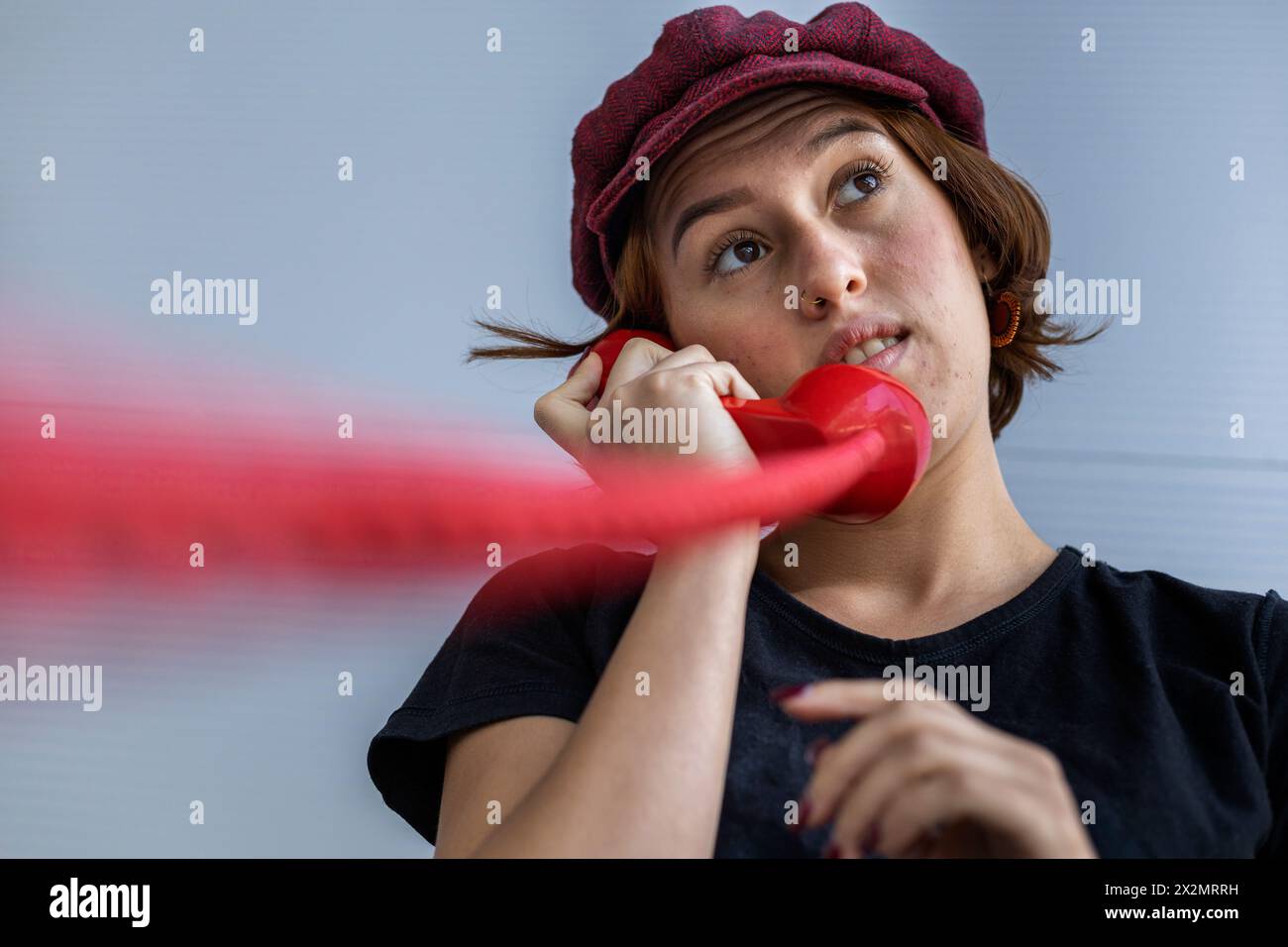 Medium short shot of Latin American young woman (22) with cap and red hair listening to conversation on retro red Handset. Low angle shot. Vintage tec Stock Photo