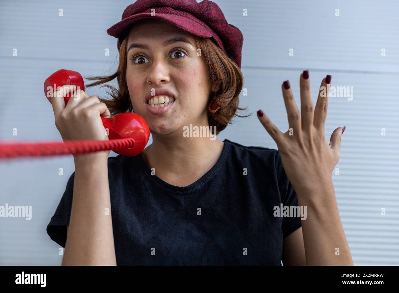 Medium short shot of young Latin American girl (22) with cap and red hair angry talking on retro red Handset. Vintage technology concept. Stock Photo