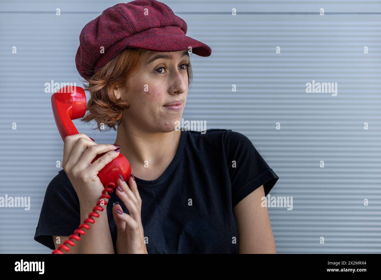 Medium short shot of young Latin American girl (22) with cap and red hair listening to a gossip on her retro red Handset and making a surprised gestur Stock Photo