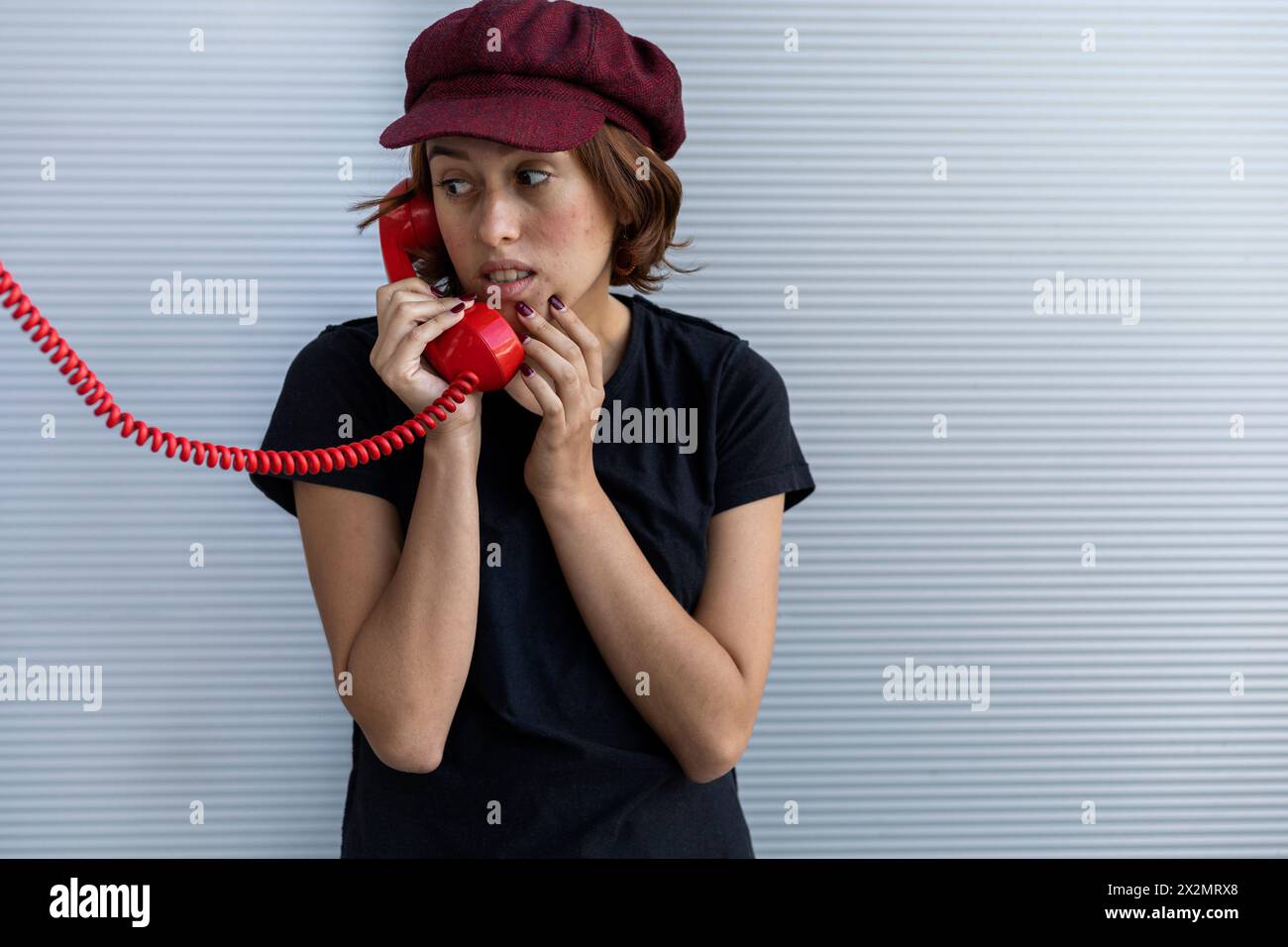 Medium short shot of a young Latin American girl (22) with a cap and red hair makes a scared face as she listens to the conversation through her retro Stock Photo