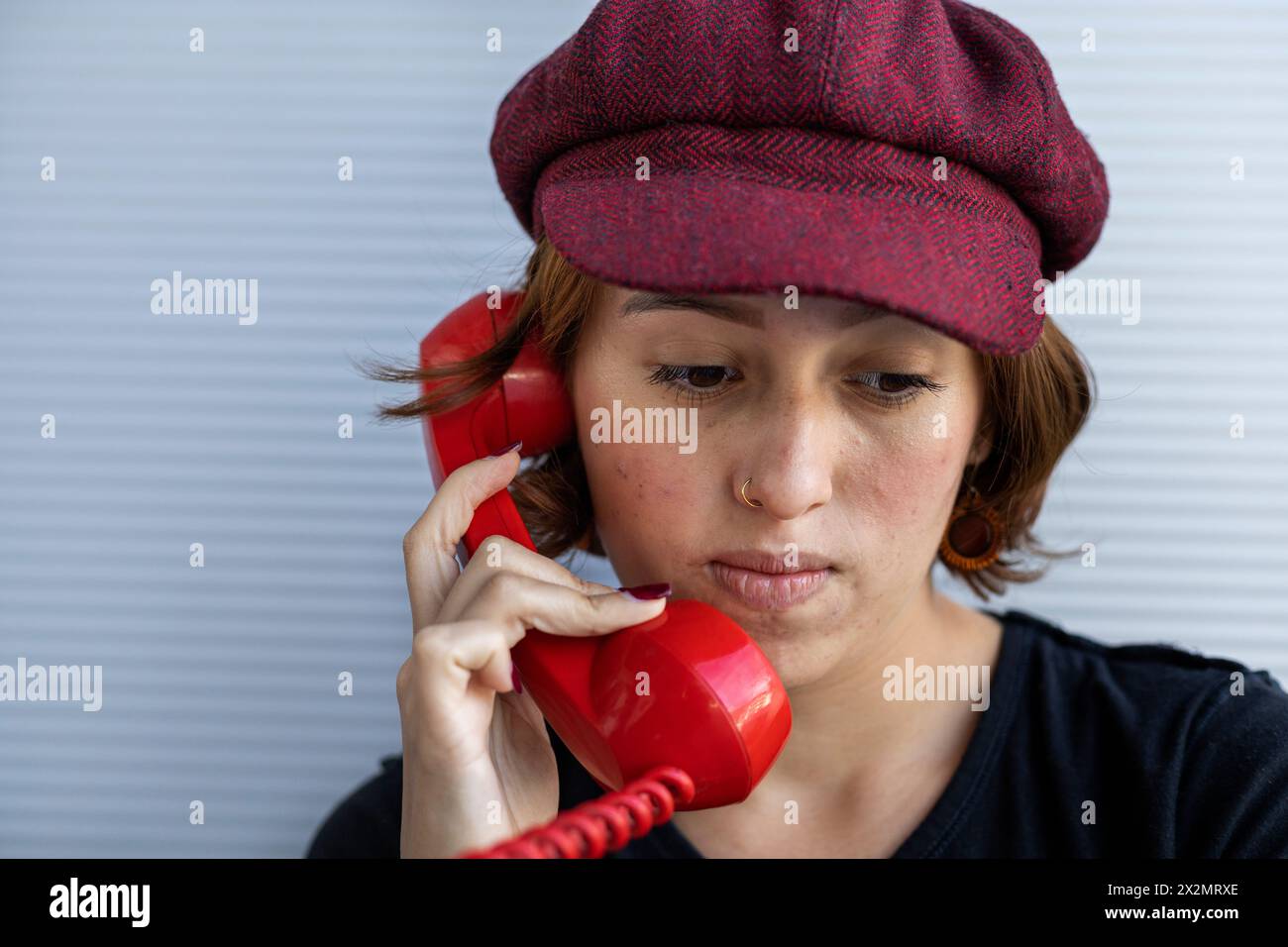 Medium short shot of Latin American young woman (22) with cap and red hair listening to conversation on retro red Handset. Low angle shot. Vintage tec Stock Photo