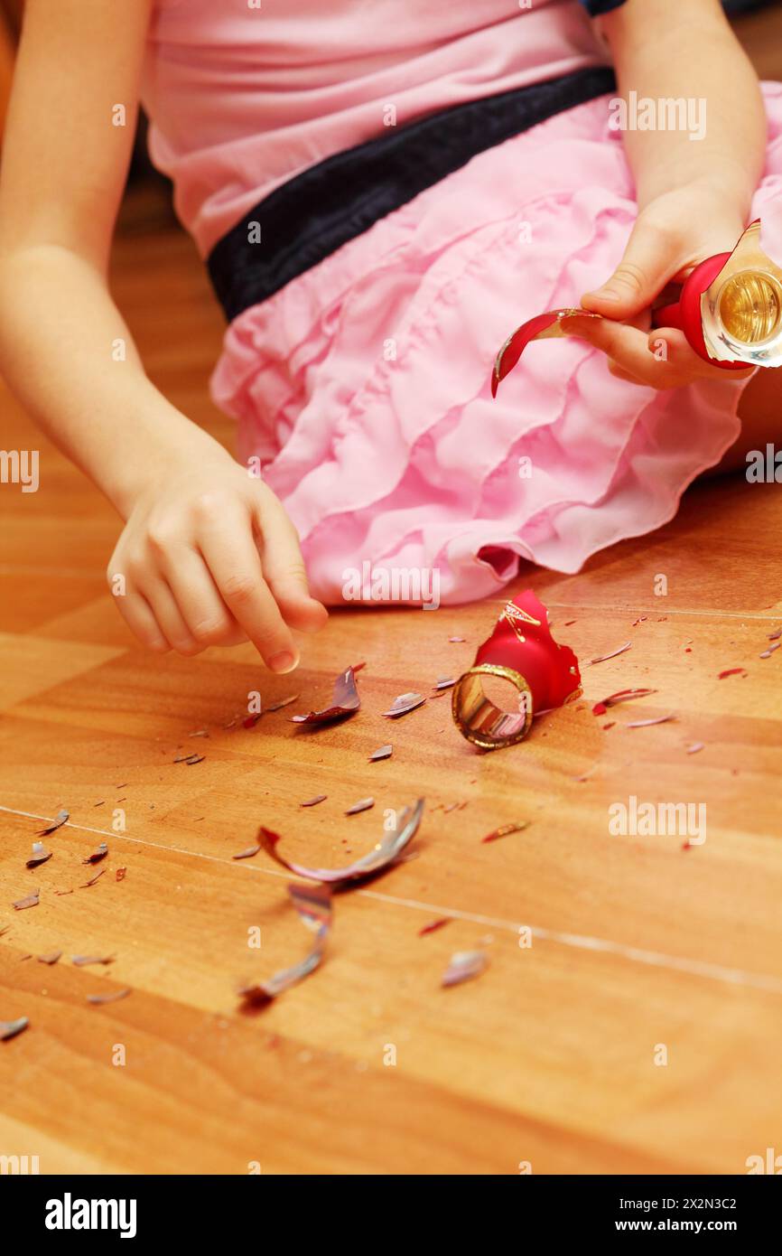 Hands of girl picking up smithereens of crushed red Christmas ball on wooden floor. Stock Photo