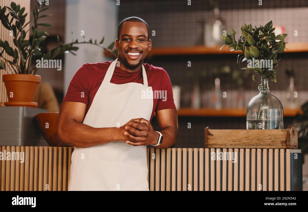 Portrait of a happy male entrepreneur standing confidently in his modern coffee shop. Man in an apron looking at the camera with a welcoming smile, em Stock Photo