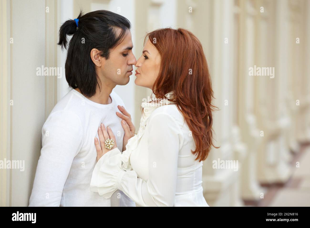 Dark-haired man and red-haired woman stand, touching noses and staring into each others eyes Stock Photo