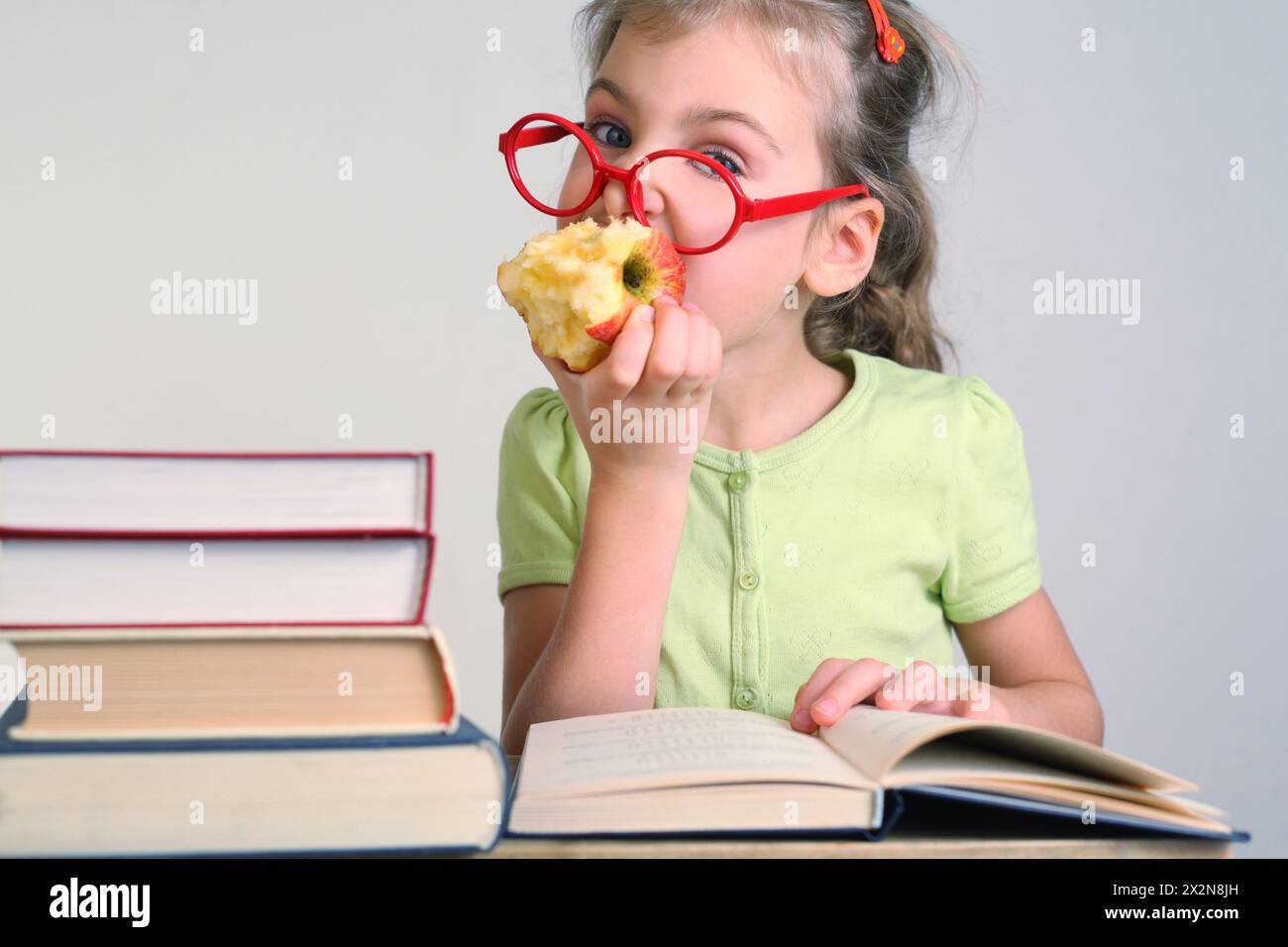 Little girl in red glasses bitten apple, seat near books Stock Photo