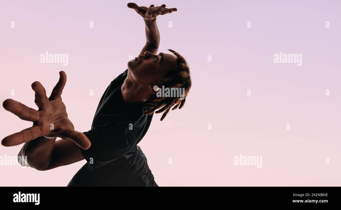 Expressive gen z male dancer with dreadlocks moves energetically in a studio, listening to music on wireless earbuds. His artistic dance moves and dyn Stock Photo